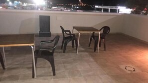 Una terraza en la azotea con una mesa y sillas, piso de baldosas y vista nocturna de la ciudad.