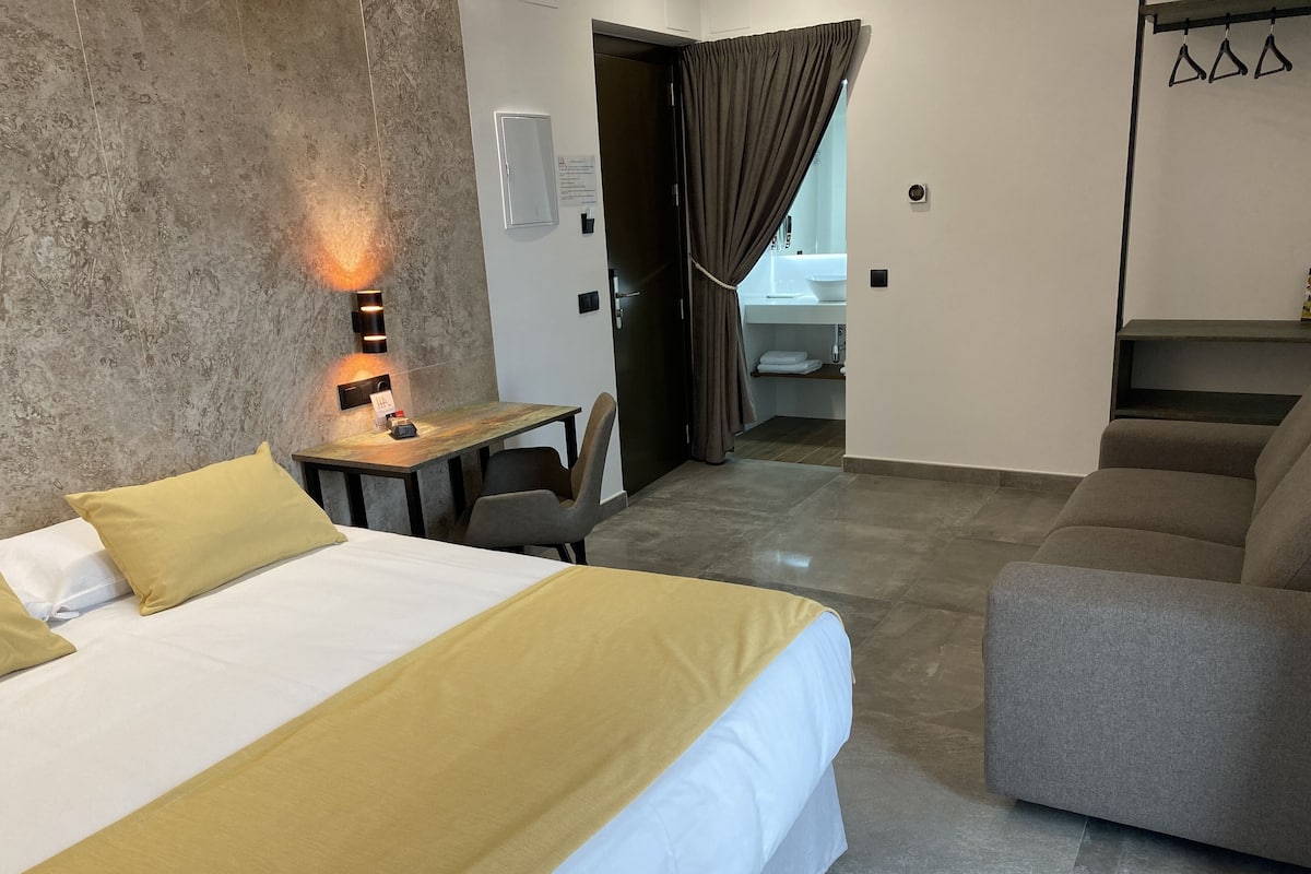 A hotel room with a bed, a desk, a chair, a sofa, and a bathroom visible through an open door.