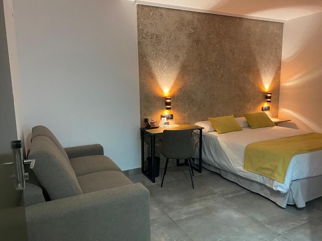 Habitación de hotel compacta con cama, escritorio y silla. Dos lámparas de pared a ambos lados de la cama.
