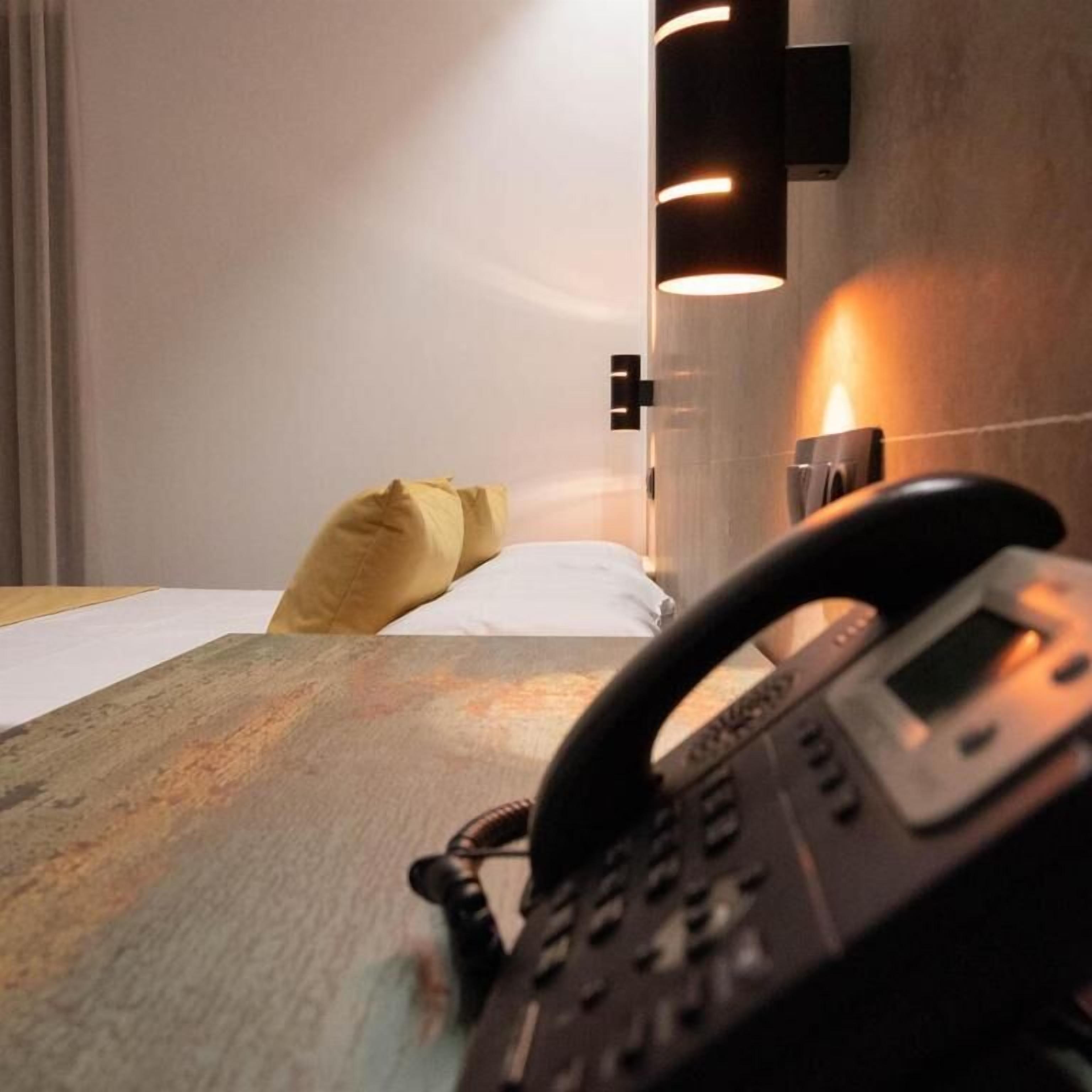 A hotel room with a bed, a bedside table, a telephone, and a wall-mounted light fixture.