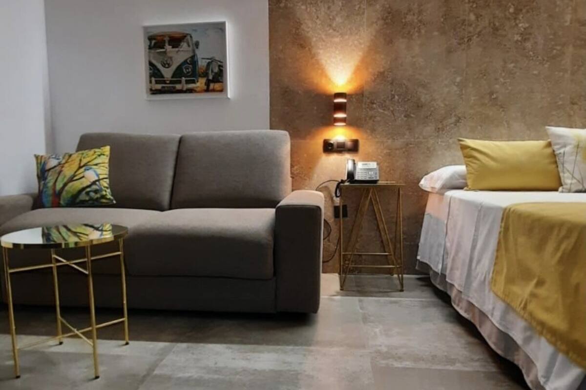 A modern room with a grey sofa, a small round table, a bed with a yellow pillow, and a textured wall.