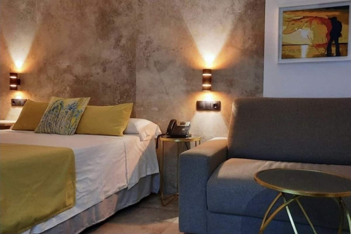 A hotel room with two beds, a sofa, a small table, and wall-mounted lamps.