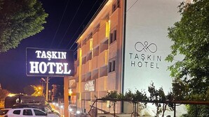 Ein Hotel mit einer großen Beschilderung, auf der "TASKIN HOTEL" steht, und einem kleineren Schild mit demselben Namen. Vor dem Gebäude ist ein weißes Auto geparkt, und das Gebäude verfügt über mehrere Stockwerke mit sichtbaren Fenstern.
