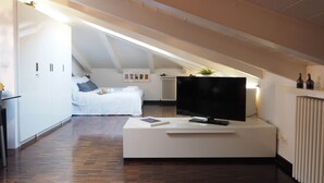 Ein modernes, helles Zimmer mit einem Flachbildfernseher auf einem weißen Standfuß, einem Bett mit weißer Bettwäsche und einem Holzboden.