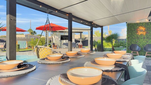Ein Essbereich im Freien mit runden Tischen und orangefarbenen Schüsseln, unter einer überdachten Terrasse mit roten Sonnenschirmen und einem Neonlichtschild.