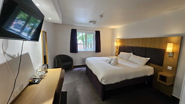 Habitación de hotel con una cama, un escritorio con computadora, una silla, un televisor montado en la pared y una ventana con cortinas.
