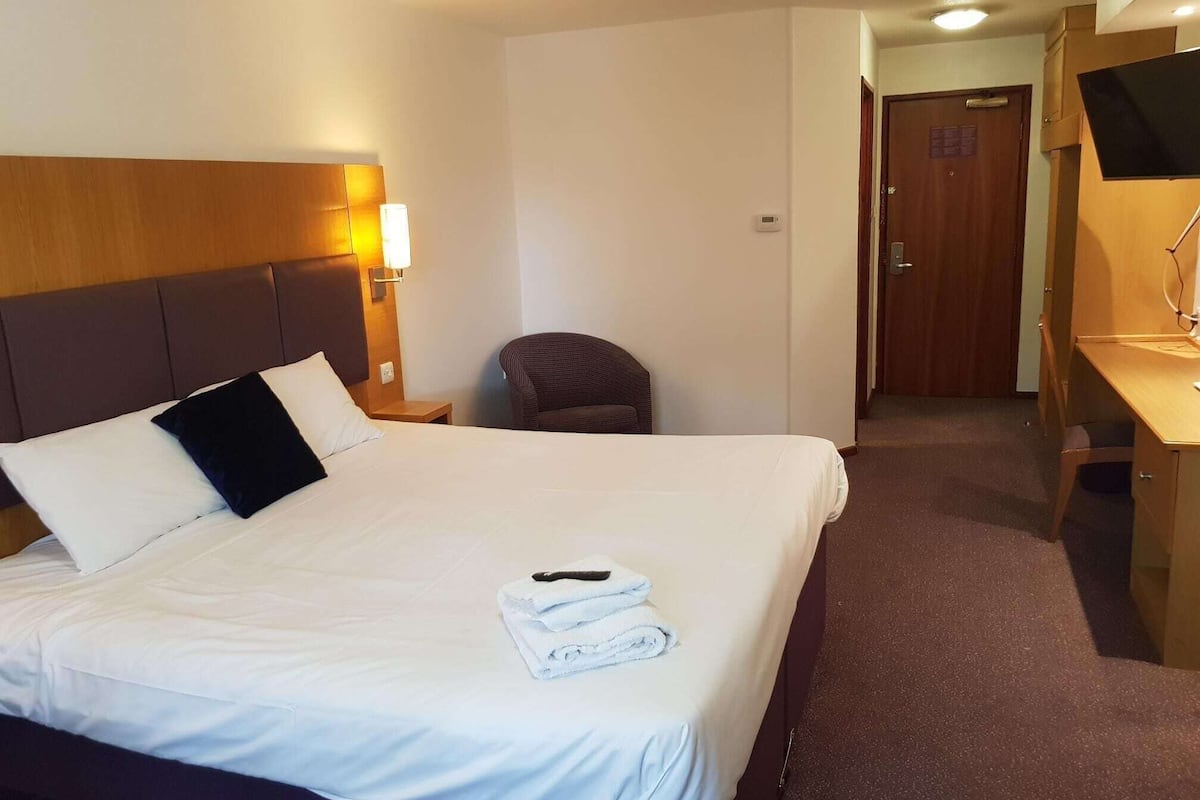 A hotel room with a large bed, a bedside table with a lamp, a chair, a desk with a computer, and a TV.