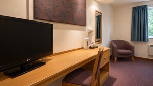 Habitación de hotel con escritorio de madera, televisor de pantalla plana, una silla, cafetera, espejo y ventana con cortinas.