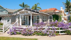 Ein zweistöckiges Haus mit überdachter Veranda, weißem Zaun und lila Blumen im Vorgarten.