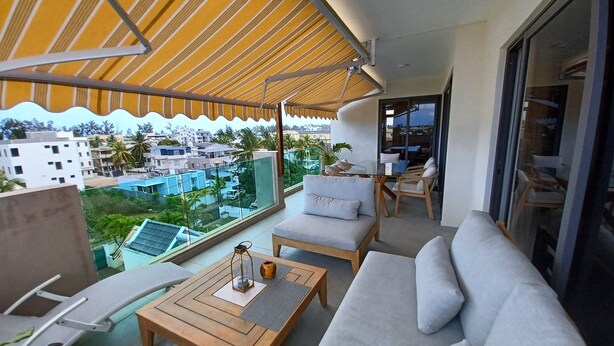 Ein Balkon mit einer Couch, Stühlen und einem Tisch, mit Blick auf ein Wohngebiet mit Gebäuden und Grünflächen.