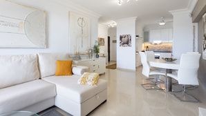 Una sala de estar moderna con un sofá blanco, una mesa de comedor con sillas blancas y una cocina al fondo.