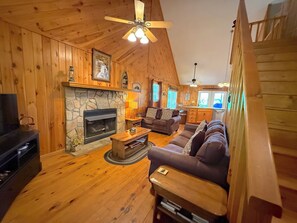 L’intérieur chaleureux d’un chalet avec une cheminée en pierre, des murs en bois, un ventilateur de plafond et un espace de vie comprenant un canapé et une table basse.