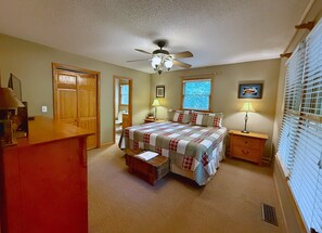 Ein Schlafzimmer mit Bett, Schreibtisch, Deckenventilator, Fenster mit Jalousien und einer Tür.