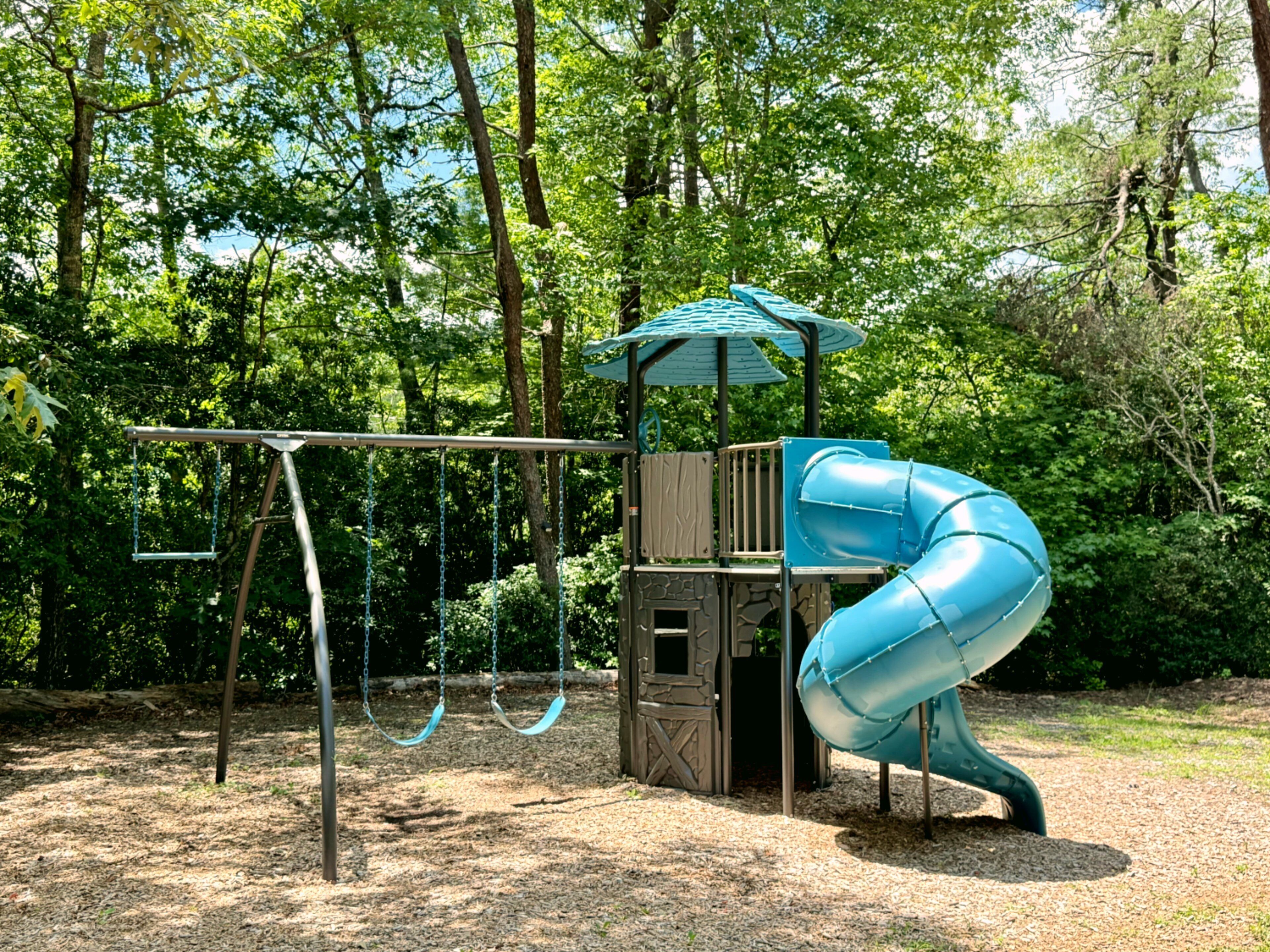 Un parque infantil con tobogán, columpios y una estructura para escalar, rodeado de árboles.
