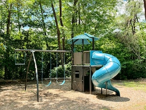 Un parque infantil con tobogán, columpios y una estructura para escalar, rodeado de árboles.