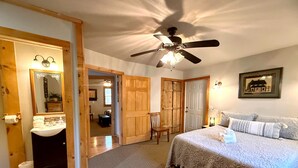 Un dormitorio con cama, ventilador de techo, baño con espejo y lavabo, y un pasillo que conduce a otra habitación.