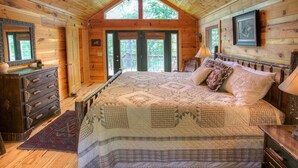 A cozy cabin bedroom with a large bed, wooden walls, and a window overlooking greenery.