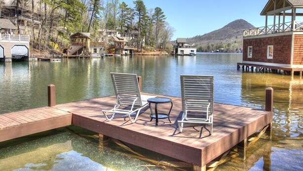 A wooden dock with two lounge chairs and a small table, overlooking a calm lake with houses and trees in the background.