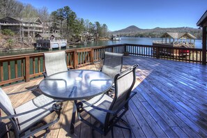 A wooden deck with a round table and four chairs, overlooking a lake with boats and houses in the background.