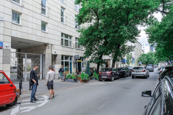 Vista de una calle con un edificio que tiene una entrada amplia, zona de mesas al aire libre con plantas y coches aparcados.