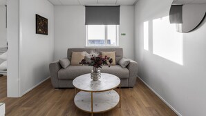A modern room with a grey sofa, a round table with a vase of flowers, and a large round mirror on the wall. - Bank Guesthouse (Reykjanesbær)