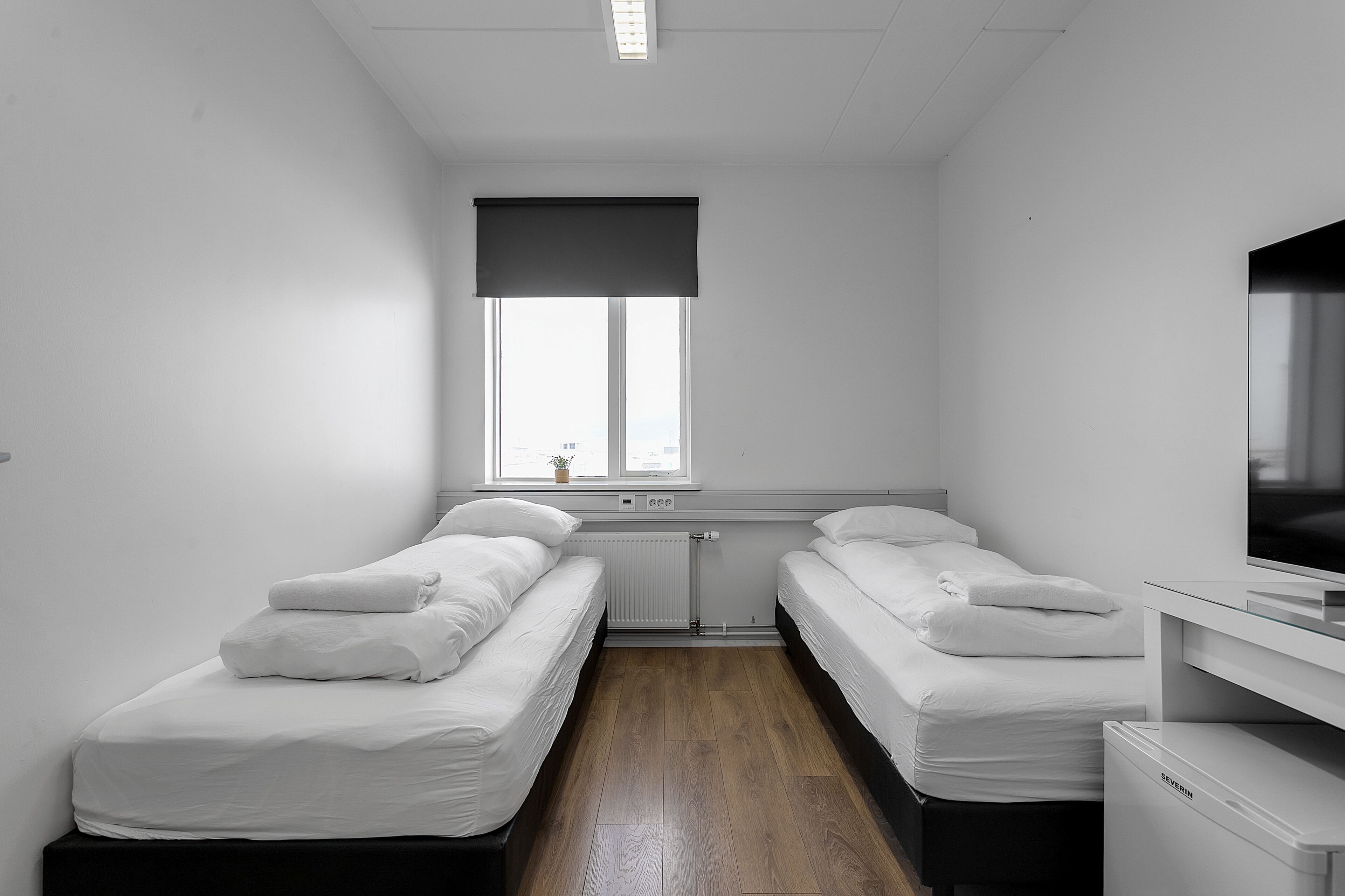 Een kleine, schone kamer met twee eenpersoonsbedden, een radiator, een raam met een donkerblinde en een tv op een standaard.