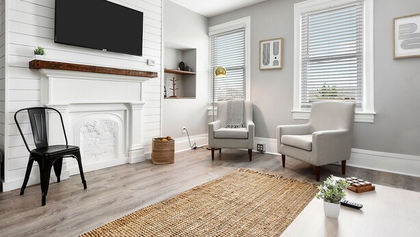 Una sala de estar con chimenea, dos sillones, un televisor montado en la pared y dos ventanas con persianas.