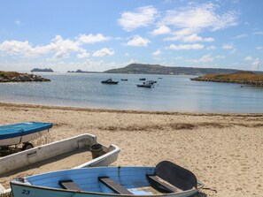 Una playa con varias embarcaciones, incluyendo una embarcación numerada y otra cubierta, cerca de aguas tranquilas, un barco lejano y colinas.