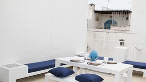 Uma área de refeições branca com almofadas azuis, uma mesa posta com pratos e um vaso azul, e vista para uma varanda com guarda-corpo.