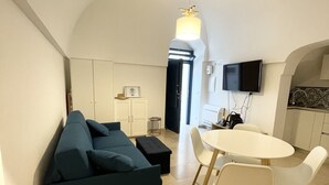 Uma sala de estar moderna com um sofá azul escuro, uma mesa de jantar redonda com cadeiras brancas e uma área de cozinha com uma TV de tela plana acima do balcão.