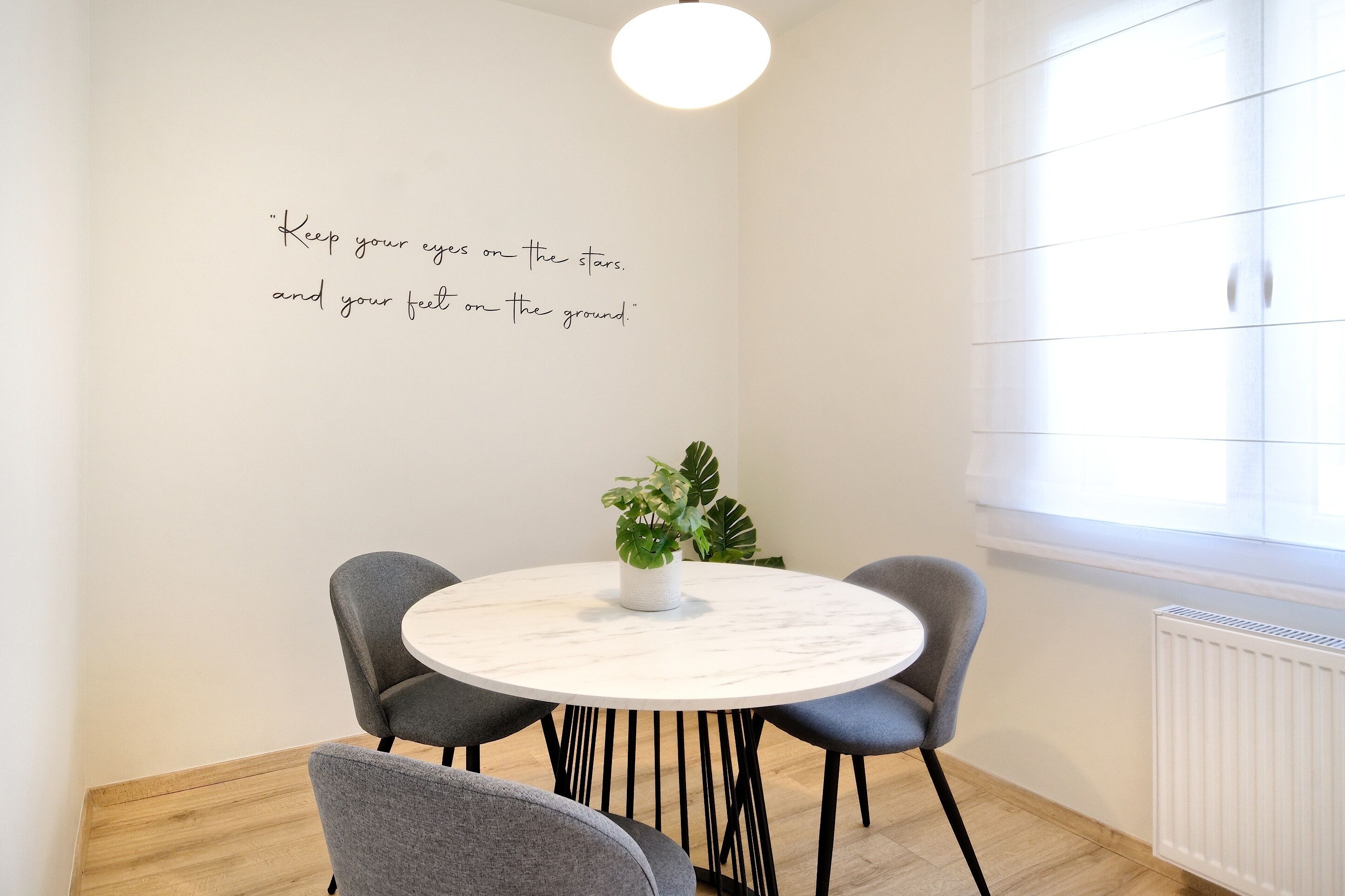 A modern dining area with a round table, four grey chairs, a potted plant, and a motivational quote on the wall.