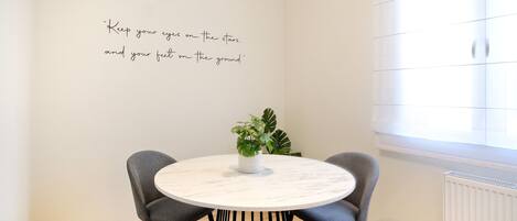 A modern dining area with a round table, four grey chairs, a potted plant, and a motivational quote on the wall.