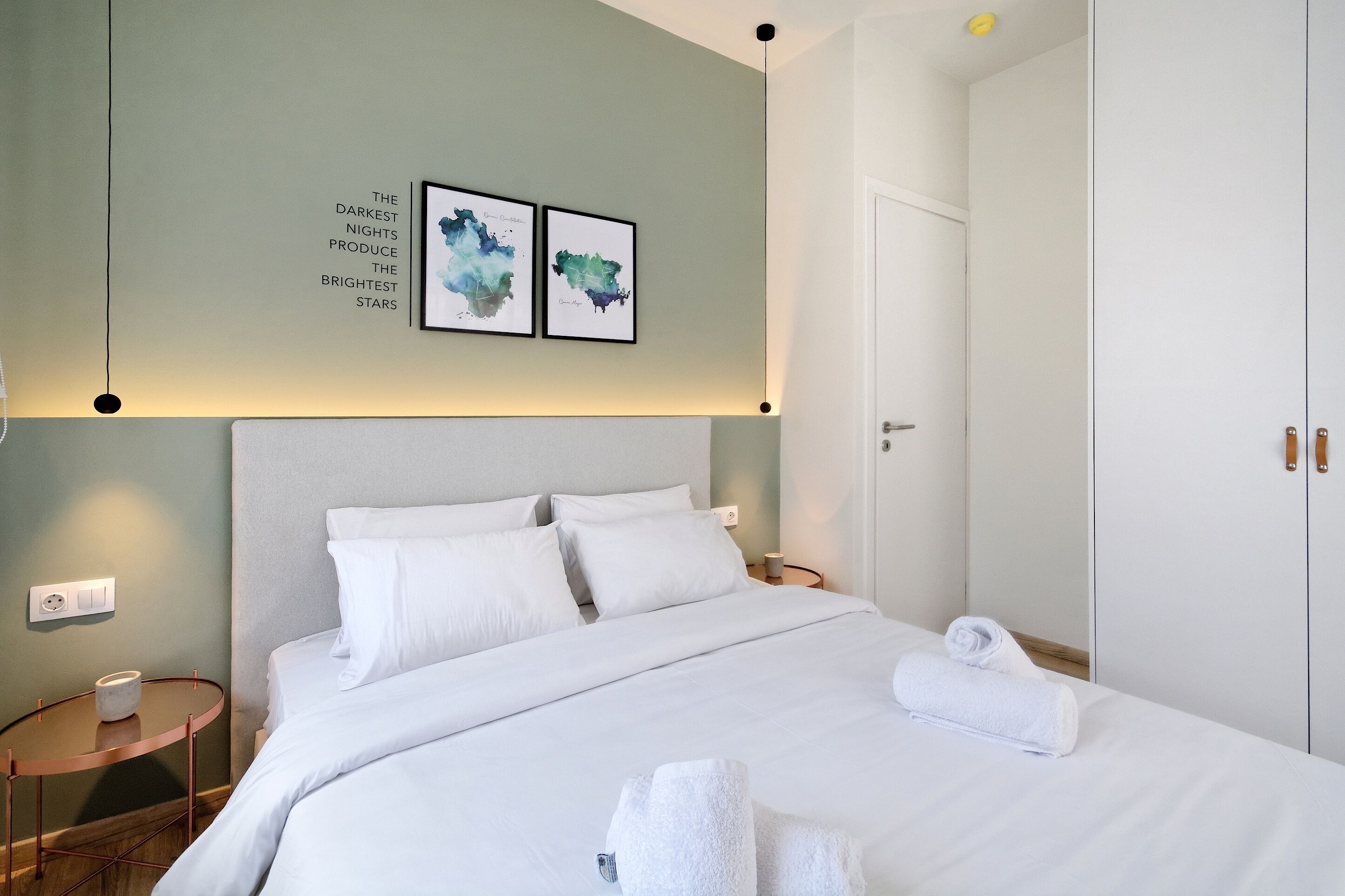 Een net opgemaakt bed met witte lakens en twee opgerolde handdoeken, een nachtkastje met een lamp, en twee ingelijste kunstwerken aan de muur.