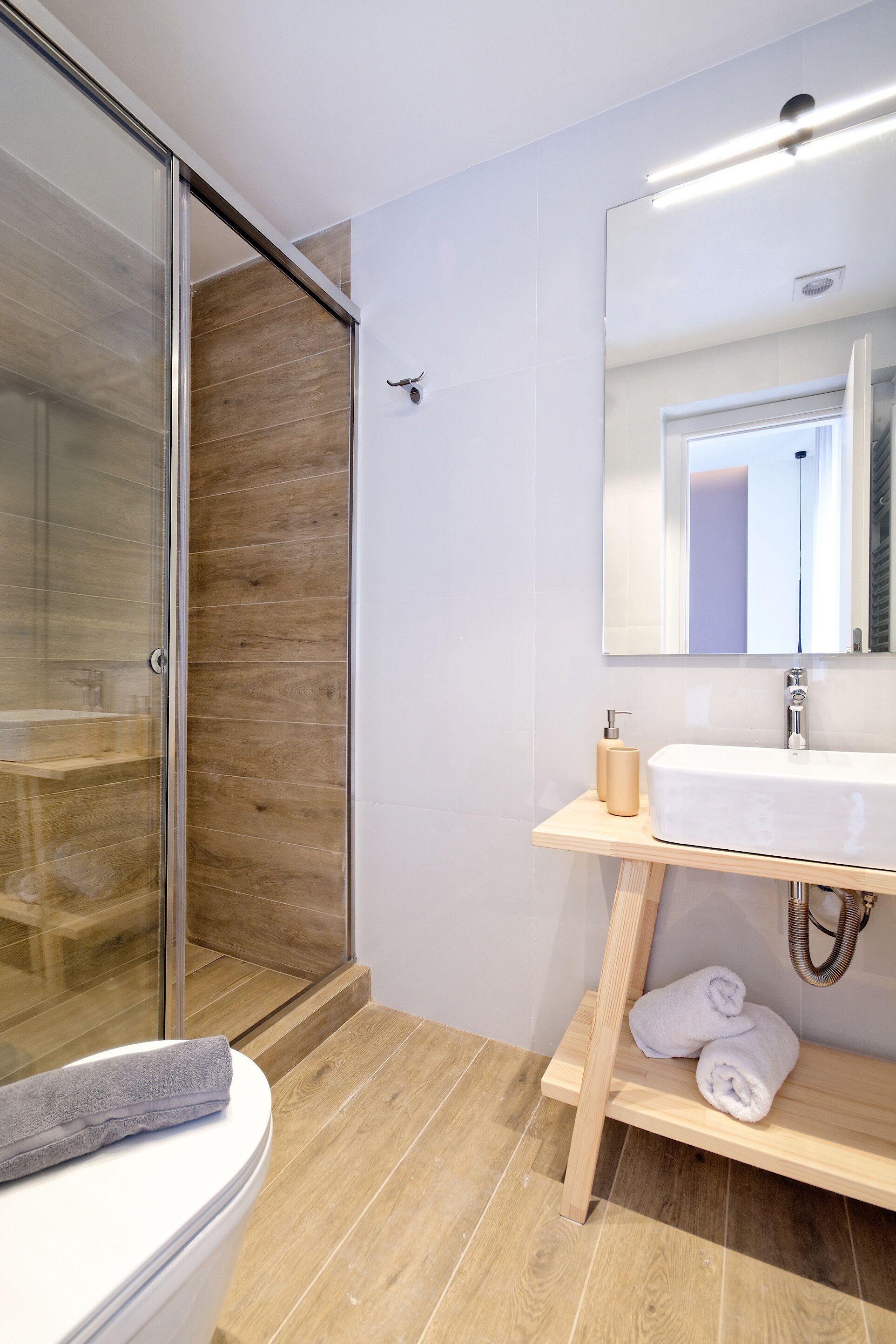 Un baño moderno con cabina de ducha de vidrio, suelo de madera, un lavabo blanco y un espejo encima.