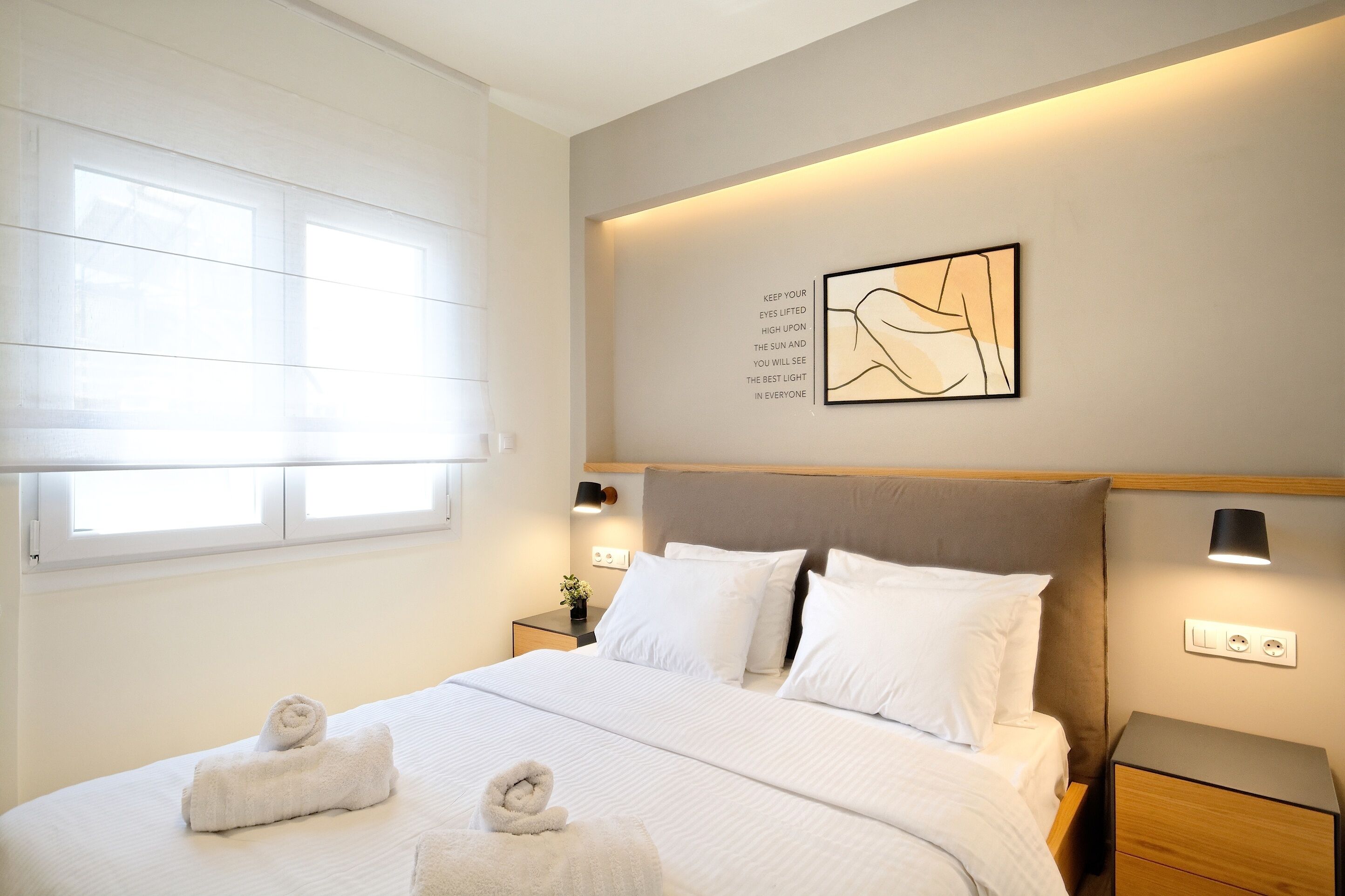 A neatly made bed with white linens and two rolled towels, a bedside table with a lamp, and a wall-mounted artwork with an inspirational quote.
