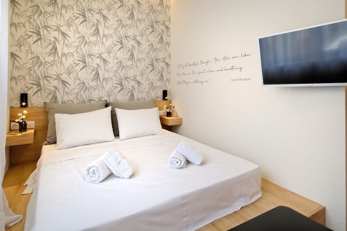 Ein ordentlich bezogenes Bett mit weißen Leinen und aufgerollten Handtüchern, eine Wand mit Bambus-Tapete und ein an der Wand befestigter Fernseher.