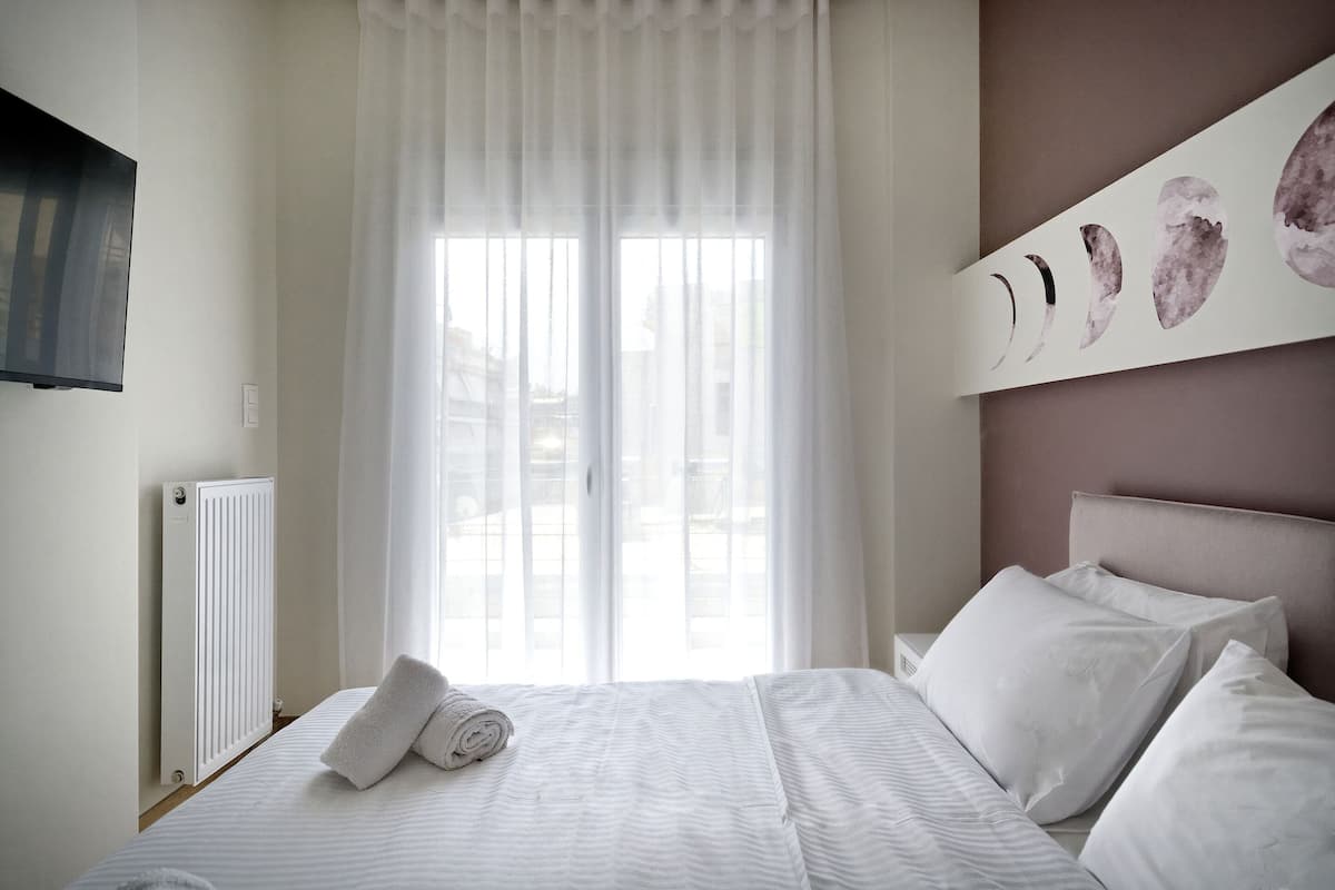 Ein Schlafzimmer mit Bett, weißen Vorhängen, einem Heizkörper und einem an der Wand befestigten Fernseher.