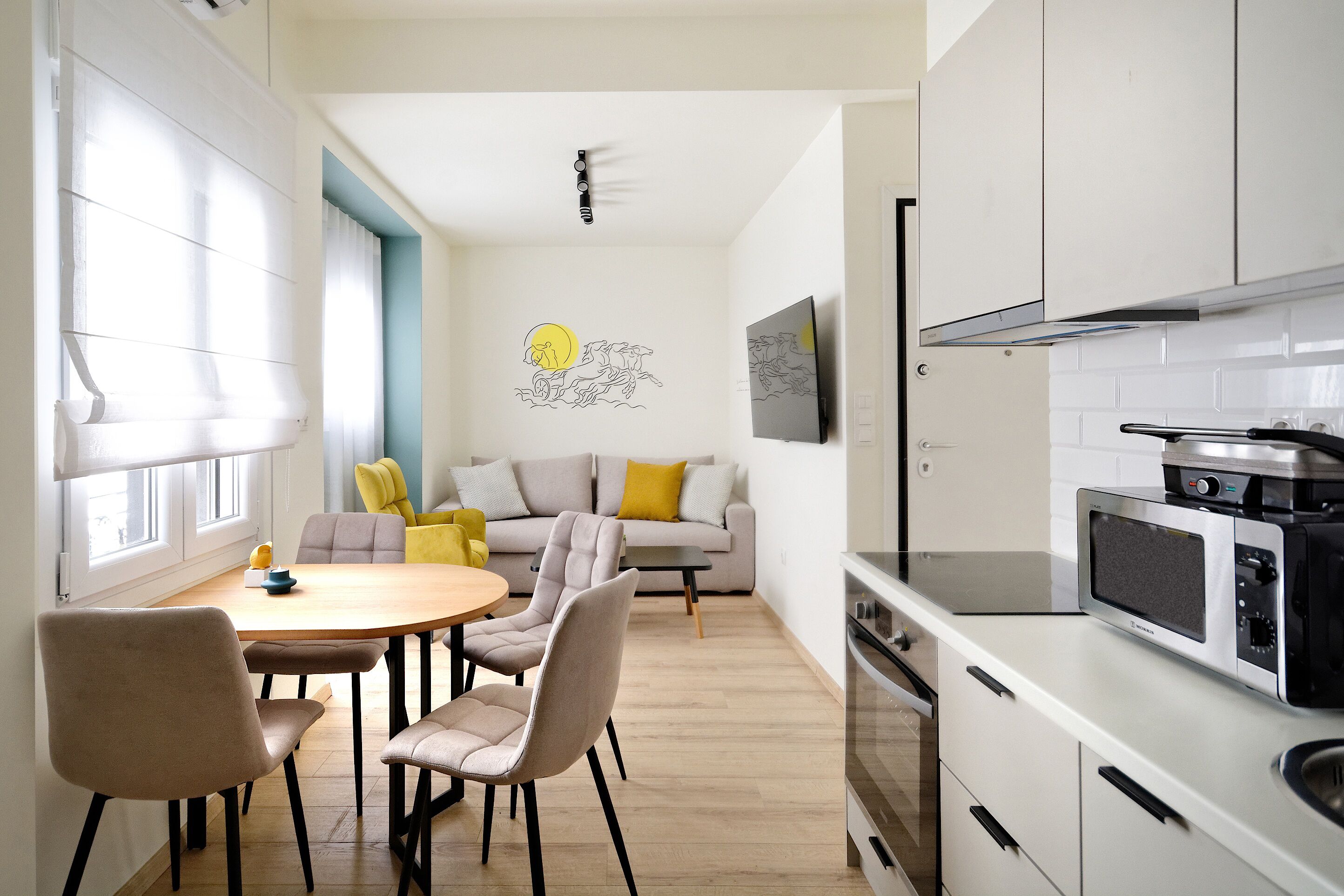 Una cocina moderna con gabinetes blancos, un microondas integrado y una estufa. Hay un comedor con una mesa redonda y sillas, y un sofá con cojines amarillos.