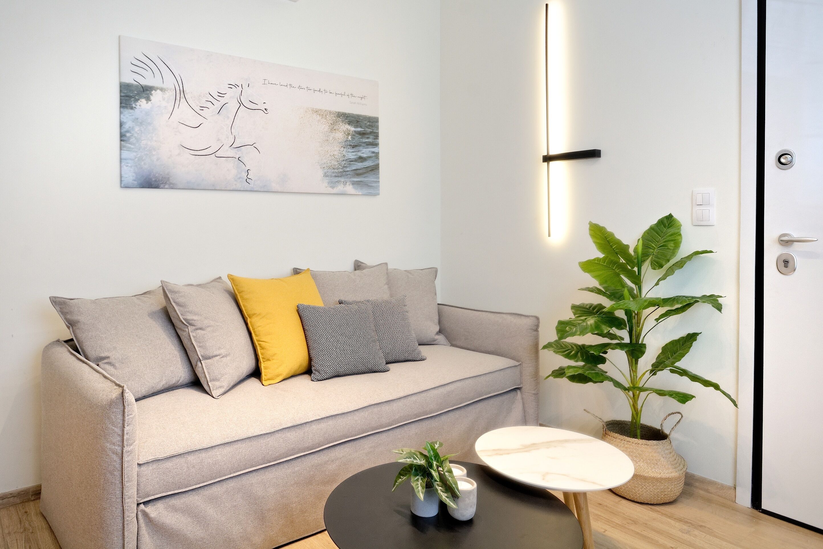 Una sala de estar moderna con un sofá gris, una almohada amarilla, una almohada gris, una mesa de centro negra, un jarrón blanco con plantas y una planta grande en una maceta tejida.
