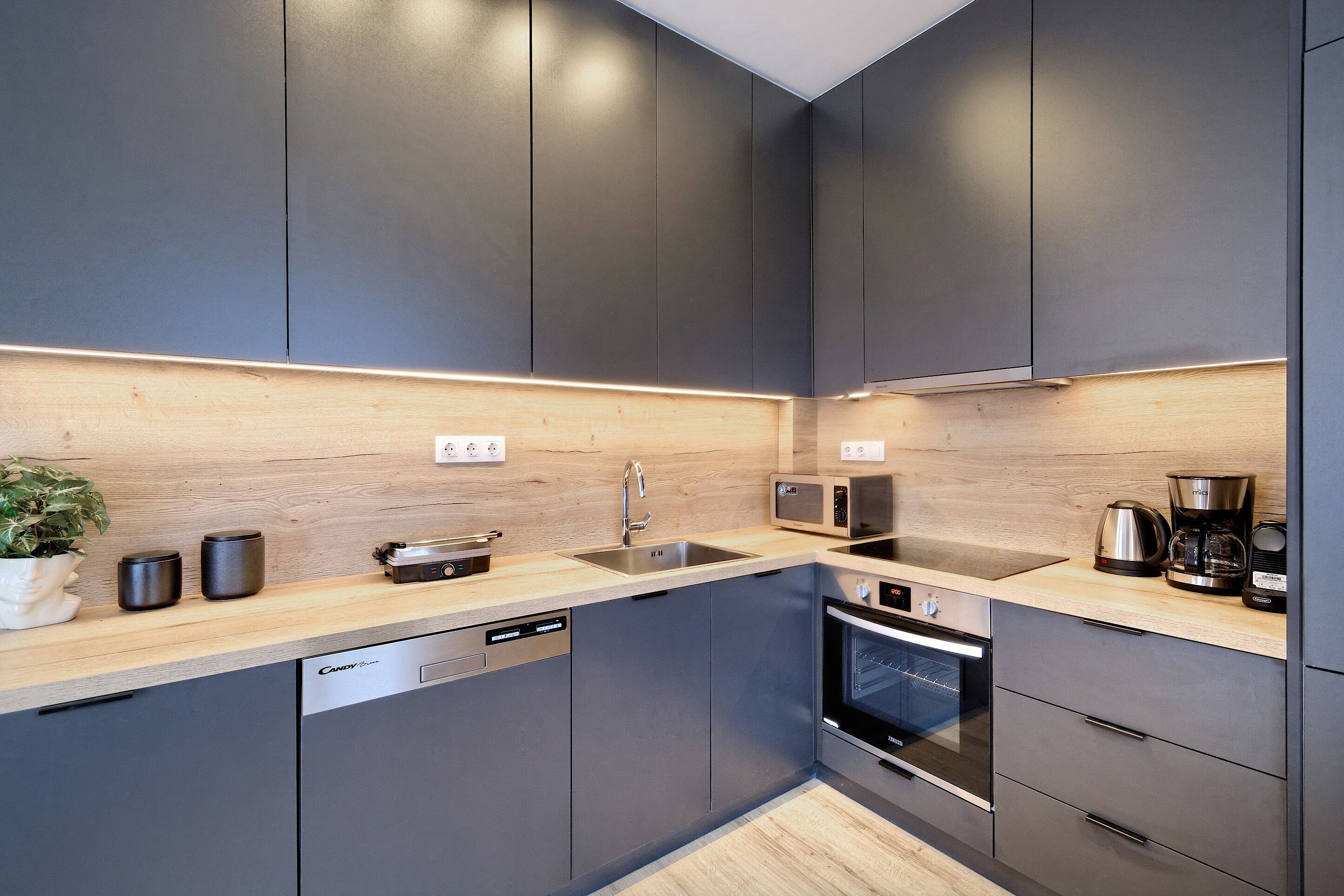 Uma cozinha moderna com armários cinza, bancada de madeira, máquina de lavar louça, forno e micro-ondas.