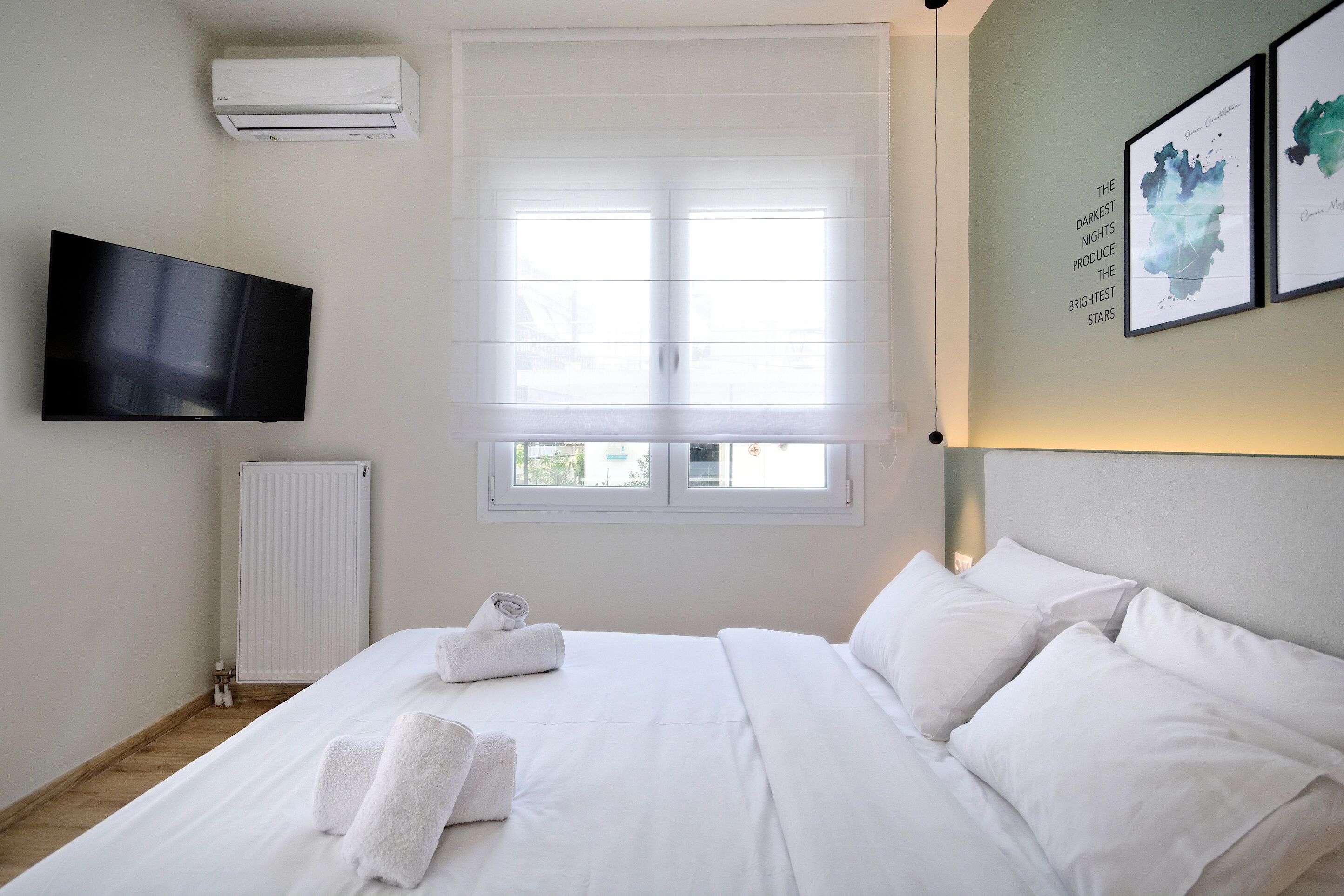 A hotel room with a bed, a flat-screen TV, a window with blinds, and a wall-mounted air conditioner.