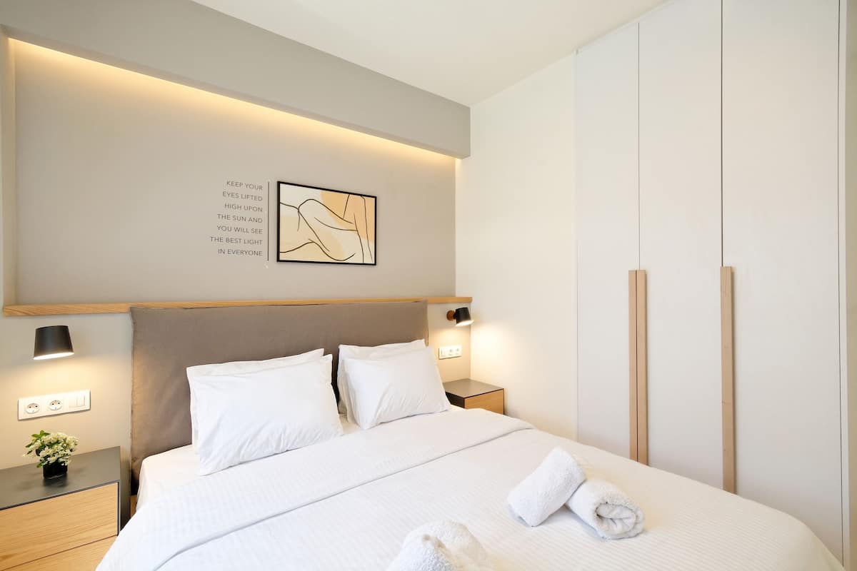 Ein modernes Hotelzimmer mit einem aufgeräumten Bett, einem Nachttisch mit Lampe, einem Wandbild und einem Kleiderschrank.