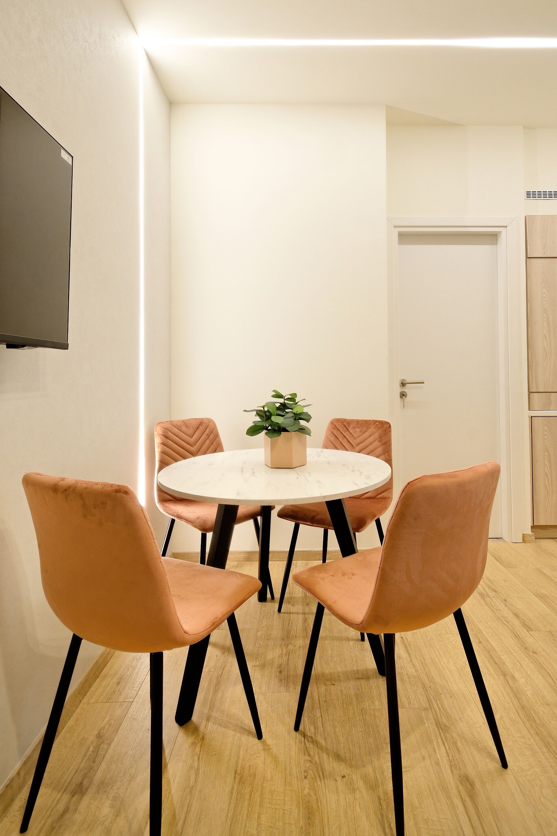 A dining area with a round table, four chairs, a potted plant, and a flat-screen TV mounted on the wall.