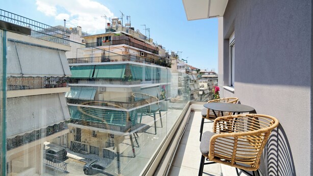 Un balcón con mobiliario de mimbre, baranda de vidrio y vistas a edificios cercanos.