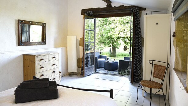 Une chambre à coucher avec un lit, une commode en bois, une chaise et une vue sur le jardin par une porte ouverte.