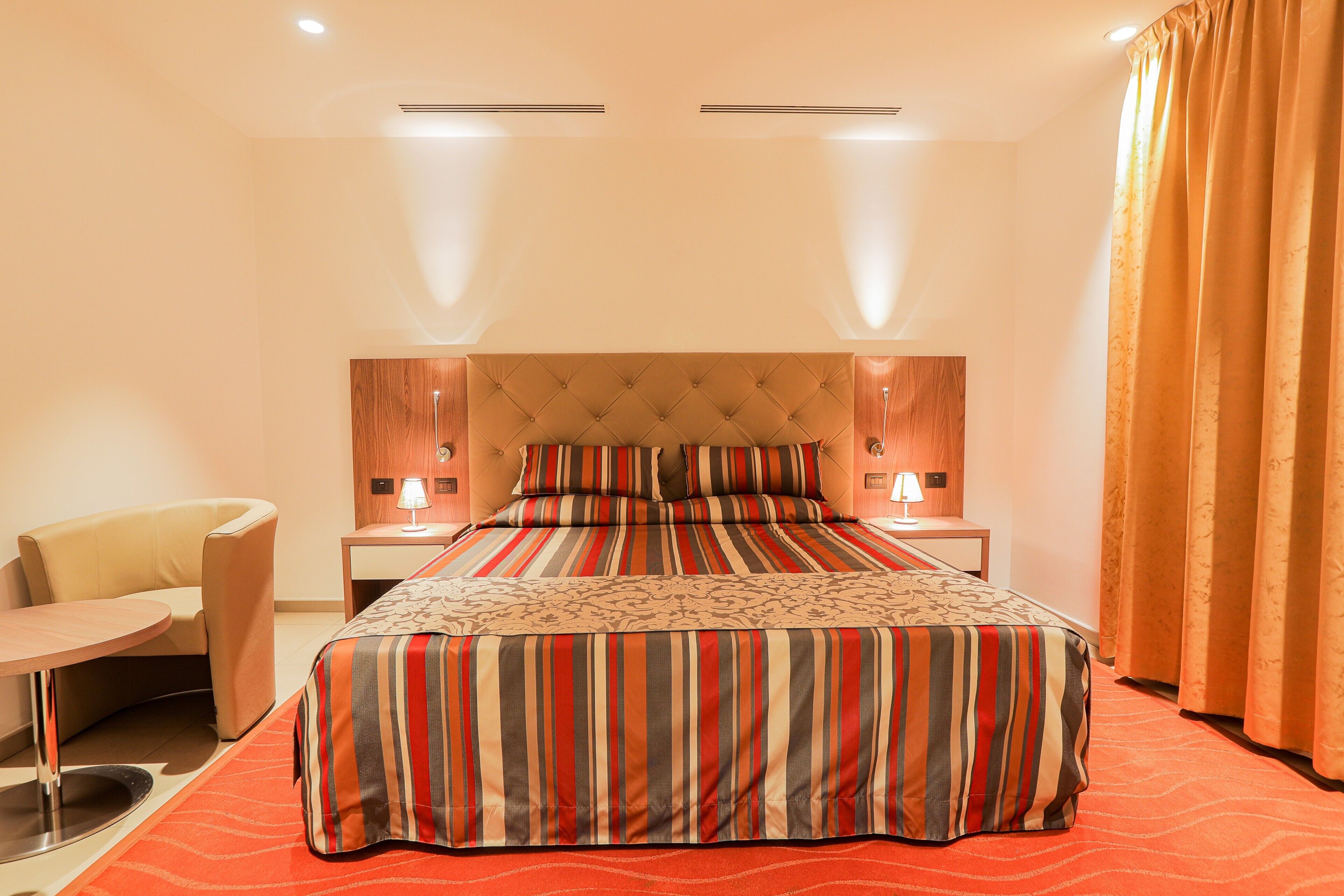 A hotel room with a large bed, a bedside table with a lamp, a chair, and a window with curtains.