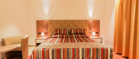 A hotel room with a large bed, a bedside table with a lamp, a chair, and a window with curtains.