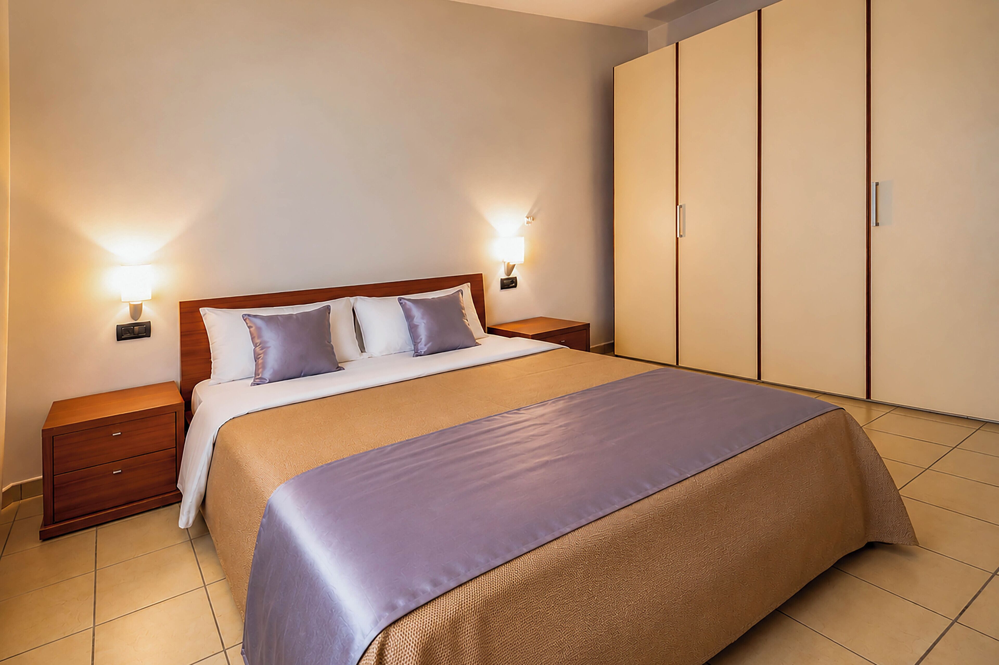 A hotel room with a large bed, two bedside tables, a wardrobe, and wall-mounted lamps.