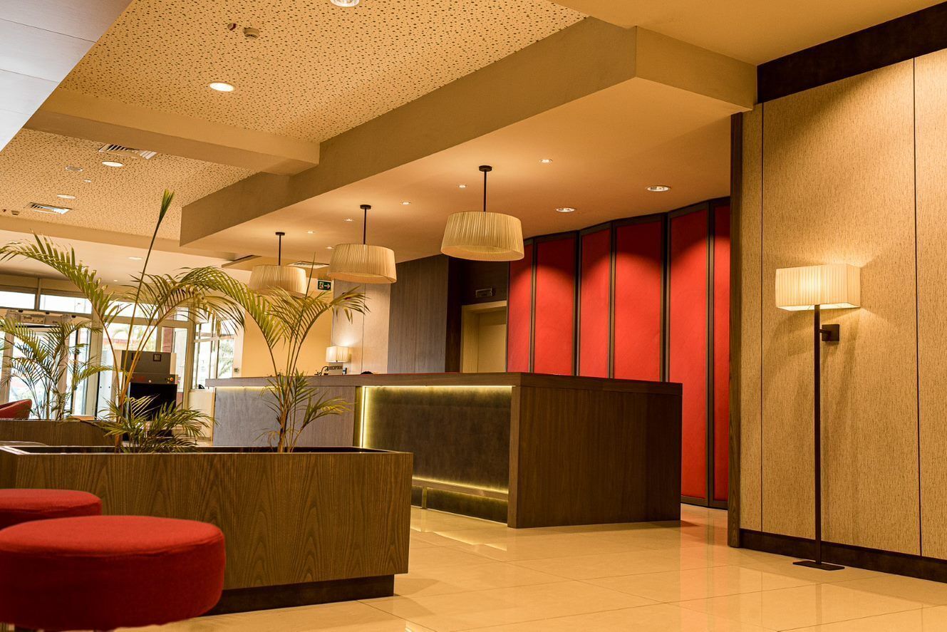 A modern hotel lobby with a reception desk, red accent walls, and a large potted plant.