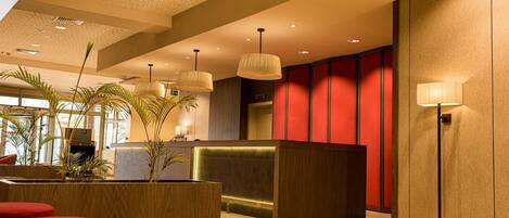 A modern hotel lobby with a reception desk, red accent walls, and a large potted plant.